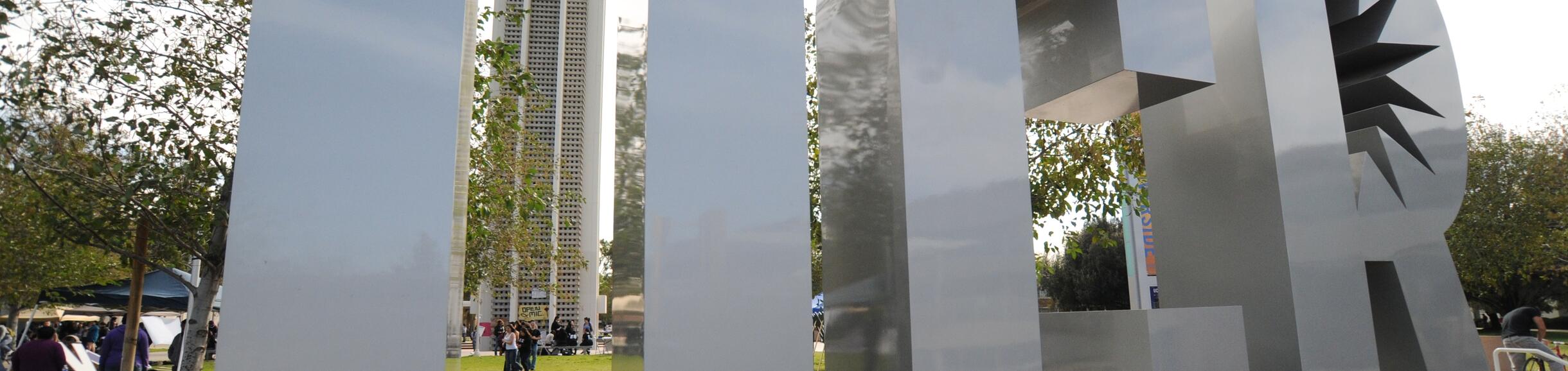Large metallic "UCR" sculpture on the University of California, Riverside campus, with the Bell Tower and trees visible in the background.  I prefer this response