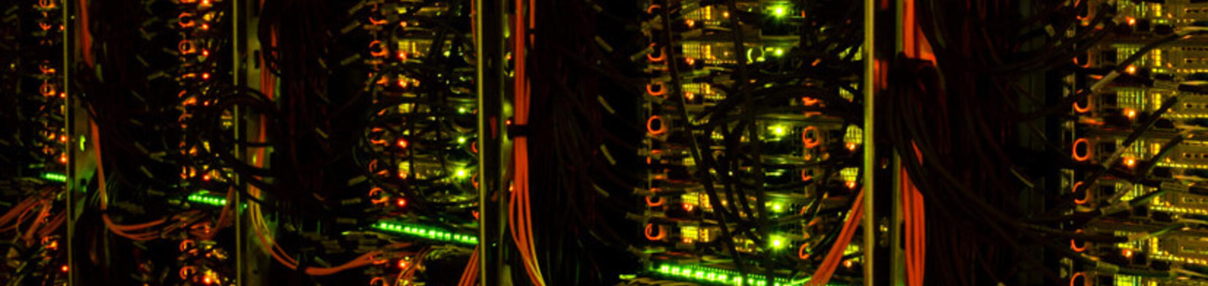 Rows of tall server racks densely packed with blinking LED lights (mostly green and orange/yellow) and a tangle of cables in a data center.