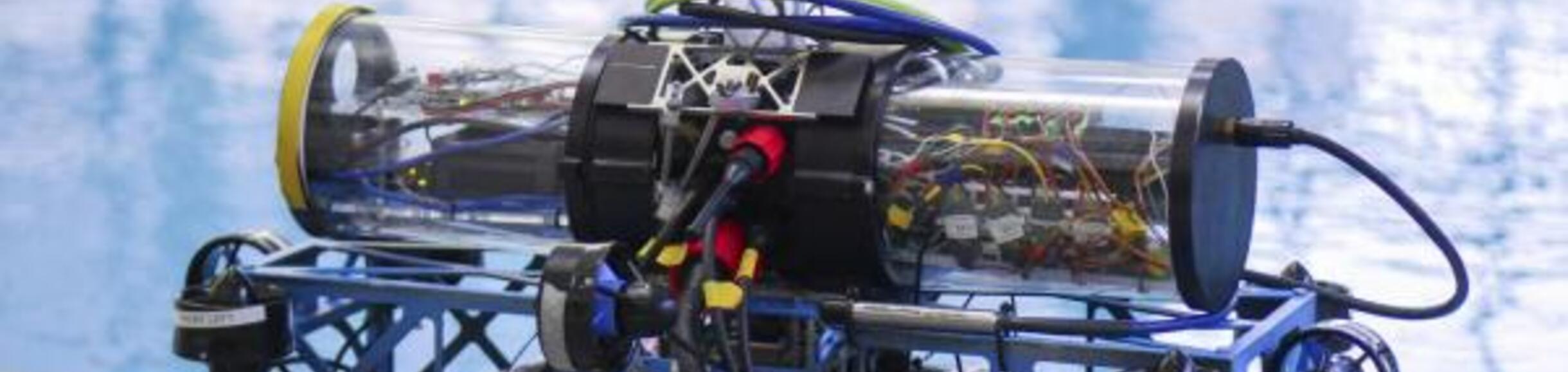 A homemade or prototype underwater remotely operated vehicle (ROV) with clear cylindrical housings revealing internal wiring, resting on the edge of a swimming pool.