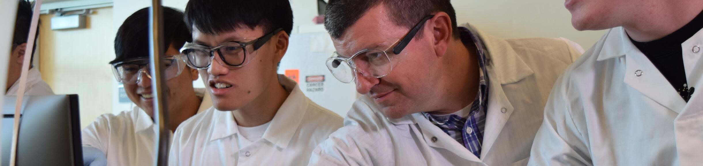 An instructor wearing safety glasses and a lab coat is guiding a group of students in protective eyewear as they look at a computer monitor in a lab setting.